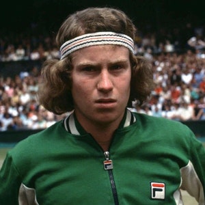 May include: A portrait of a tennis player wearing a green zip-up jacket with white trim and a white headband with red and black stripes. The jacket features a FILA logo. The player has light brown hair and is looking directly at the camera.