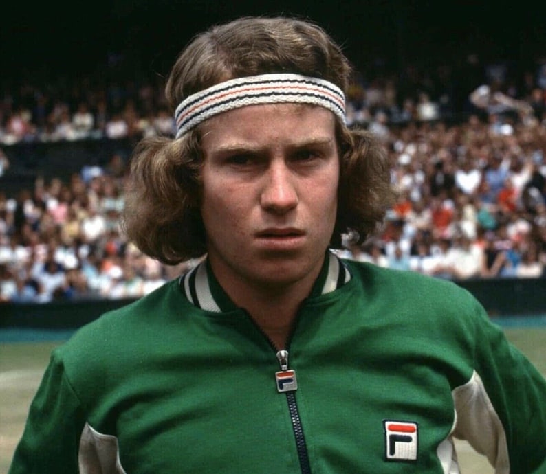 May include: A portrait of a tennis player wearing a green zip-up jacket with white trim and a white headband with red and black stripes. The jacket features a FILA logo. The player has light brown hair and is looking directly at the camera.
