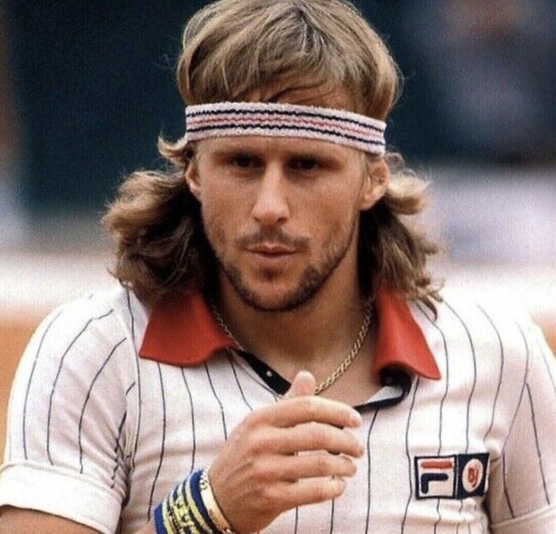 May include: A man wearing a white tennis shirt with vertical blue stripes, a red collar, and a Fila logo. He has a white headband with red and blue stripes, a gold chain, and a wristband. The man has blond hair and a beard.