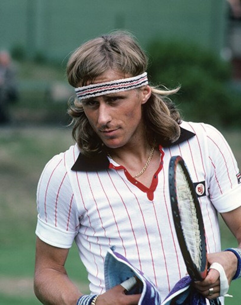 May include: A vintage tennis player wearing a white shirt with red vertical stripes and a black collar. He is holding a tennis racket and a towel. A white headband with red and black stripes is on his head.