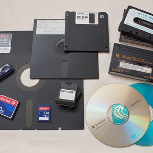 Pode incluir: Uma cole&ccedil;&atilde;o de v&aacute;rios meios de armazenamento, incluindo disquetes, fitas cassete, CD-ROM e cart&otilde;es de mem&oacute;ria. Os meios est&atilde;o dispostos sobre uma superf&iacute;cie branca. Os disquetes s&atilde;o pretos e est&atilde;o etiquetados com "TDK MF-2HD" e "ID". As fitas cassete s&atilde;o pretas e est&atilde;o etiquetadas com "JET SET WILLY II FOR THE SINCLAIR SPECTRUM" e "8mm data cartridge". Os CD-ROM s&atilde;o prateados e est&atilde;o etiquetados com "Verbatim" e "TDK CD-R80". Os cart&otilde;es de mem&oacute;ria s&atilde;o azuis e est&atilde;o etiquetados com "SanDisk 2.0" e "SanDisk 16".