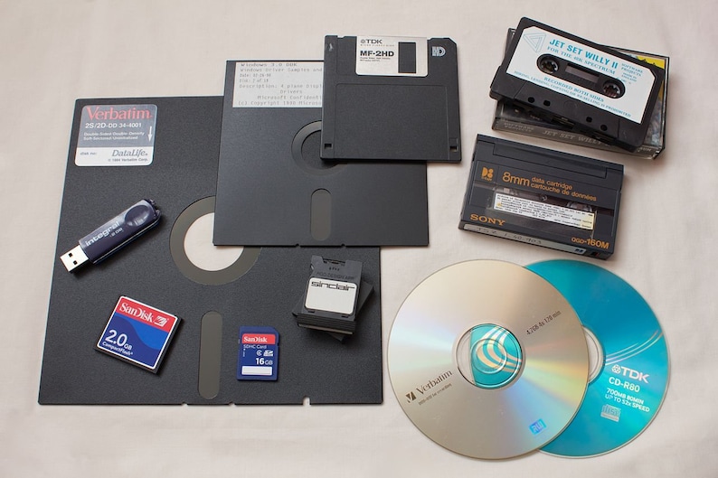 Pode incluir: Uma cole&ccedil;&atilde;o de v&aacute;rios meios de armazenamento, incluindo disquetes, fitas cassete, CD-ROM e cart&otilde;es de mem&oacute;ria. Os meios est&atilde;o dispostos sobre uma superf&iacute;cie branca. Os disquetes s&atilde;o pretos e est&atilde;o etiquetados com "TDK MF-2HD" e "ID". As fitas cassete s&atilde;o pretas e est&atilde;o etiquetadas com "JET SET WILLY II FOR THE SINCLAIR SPECTRUM" e "8mm data cartridge". Os CD-ROM s&atilde;o prateados e est&atilde;o etiquetados com "Verbatim" e "TDK CD-R80". Os cart&otilde;es de mem&oacute;ria s&atilde;o azuis e est&atilde;o etiquetados com "SanDisk 2.0" e "SanDisk 16".