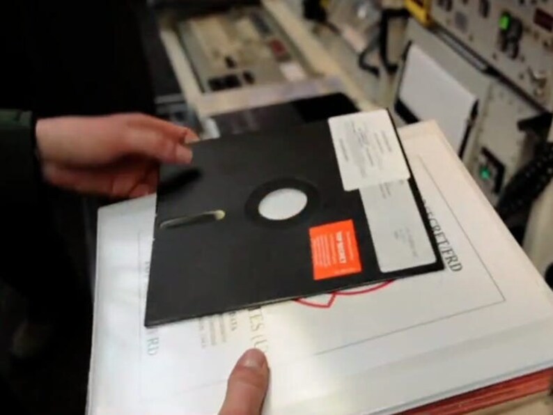 Pode incluir: Um disquete preto com um r&oacute;tulo vermelho que diz "3M" est&aacute; sendo segurado por uma pessoa. O disco est&aacute; em cima de um documento branco com o texto "GEHEIMTIERD" impresso nele.