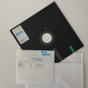 Pode incluir: Um disquete preto com um r&oacute;tulo que diz "diskettes" e uma aba verde. Um peda&ccedil;o de papel branco com o texto "[302,1] BINFILES 7600 Binary files From MBACAD Directory By (302,11 25-JUL-84 14:20 POREMPLOY, BIN 98. 25-JUL-64 14:17 POZDRI, BIN 141. 25-JUL-14 14:18 BOOP IC, BIN 70. 14:19 Total of 391/391. blocks in 4. files" est&aacute; em cima do disco.