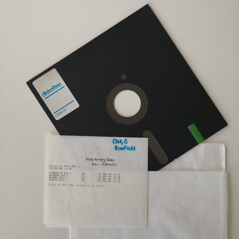 Pode incluir: Um disquete preto com um r&oacute;tulo que diz "diskettes" e uma aba verde. Um peda&ccedil;o de papel branco com o texto "[302,1] BINFILES 7600 Binary files From MBACAD Directory By (302,11 25-JUL-84 14:20 POREMPLOY, BIN 98. 25-JUL-64 14:17 POZDRI, BIN 141. 25-JUL-14 14:18 BOOP IC, BIN 70. 14:19 Total of 391/391. blocks in 4. files" est&aacute; em cima do disco.