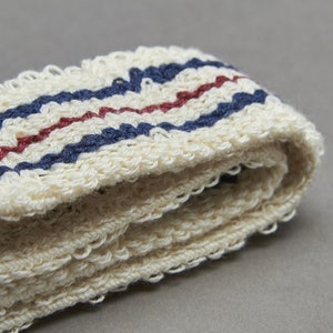 May include: Close-up of a folded, off-white, textured fabric with horizontal stripes. The stripes are in navy blue and burgundy. The fabric appears to be a towel or washcloth.