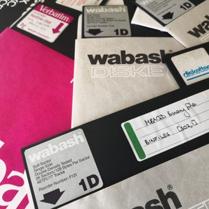 Pode incluir: Close-up de uma pilha de disquetes com a etiqueta "wabash" com o texto "Soft Sector Single Side Double Density Tested 26 Sectors/128 Bytes Per Sector 48TPI/77 Tracks Reorder Number: F131 1D" e "MBACD Binary Files BINFILES [302,7]"