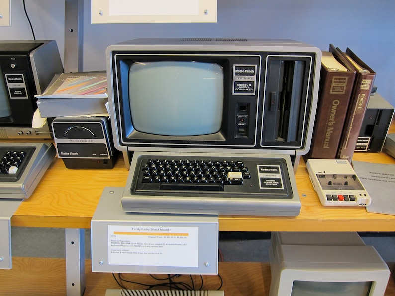 Pode incluir: Um computador vintage Radio Shack TRS-80 Modelo III com um monitor preto e branco, um teclado, uma unidade de disquete e uma unidade de fita cassete. O computador est&aacute; em cima de uma mesa de madeira com uma etiqueta que diz "TRS-80 Radio Shack Model III".