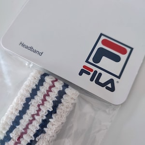 May include: White headband with blue and maroon stripes, packaged with a white card. The card features the word "Headband" and the Fila logo in navy, red, and white.