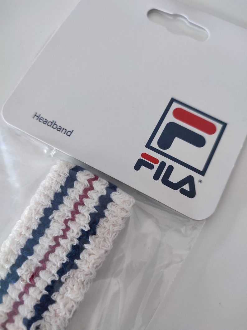 May include: White headband with blue and maroon stripes, packaged with a white card. The card features the word "Headband" and the Fila logo in navy, red, and white.