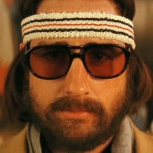 Può includere: Un uomo con la barba e capelli lunghi e castani indossa una fascia bianca con strisce nere e occhiali da sole marroni. Sta guardando direttamente la telecamera.