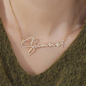 Puede incluir: Un collar de cadena dorada con un colgante de placa de nombre cursivo que dice "Summer" en diamantes de imitación brillantes.