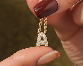 May include: A gold chain necklace with a letter A pendant encrusted with small clear stones. The necklace is being held by a hand with red nail polish.
