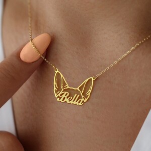 May include: A gold-colored necklace featuring a stylized dog face with the name "Bella" cut out in script. The pendant hangs from a delicate chain. The necklace is worn around the neck.