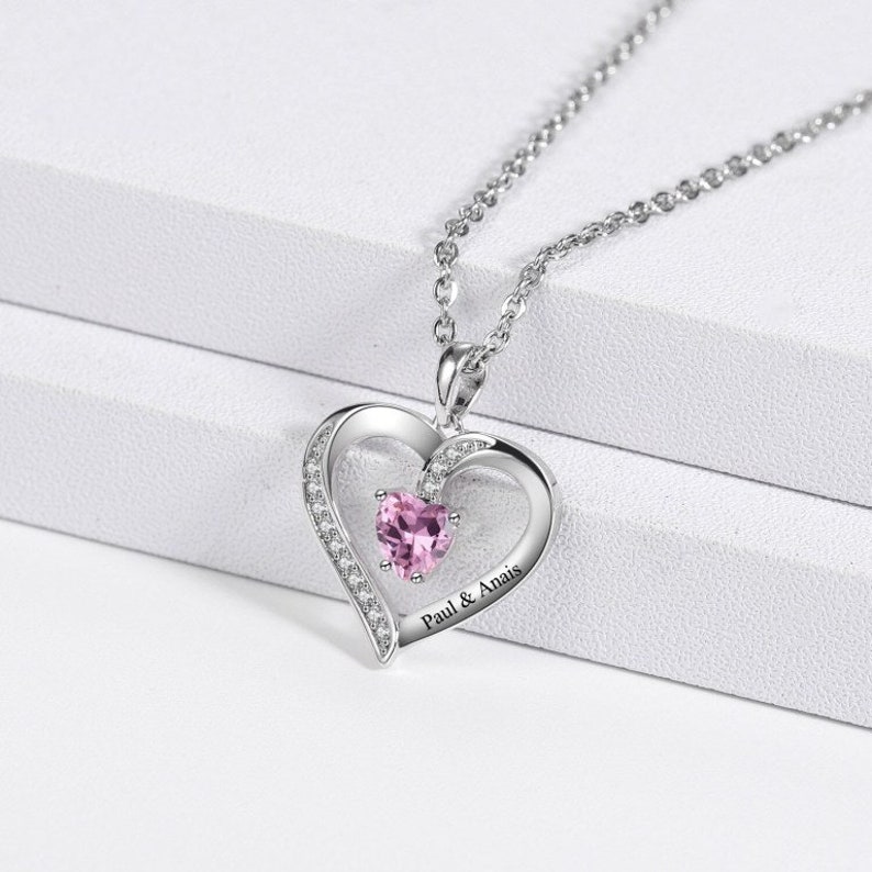 May include: A silver necklace with a heart-shaped pendant. The pendant features a smaller heart in the centre with a pink gemstone. The pendant is engraved with the text "Paul & Anais".