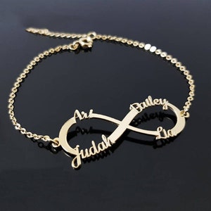 May include: A gold-coloured infinity bracelet with the names Ari, Judah, Bailey, and Lia. The bracelet has a delicate chain and a polished finish. The names are in a cursive font.