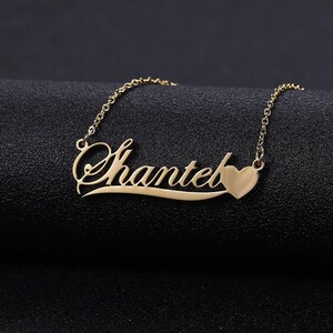 Puede incluir: Un collar dorado con el nombre "Chantel" en fuente cursiva. Una pequeña forma de corazón está al final del nombre. El collar está sobre una superficie negra.