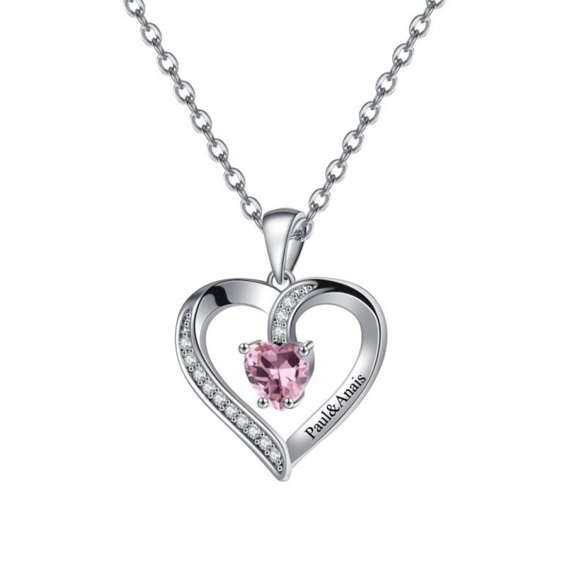 May include: Silver necklace featuring a heart-shaped pendant. The pendant has a smaller heart-shaped pink gemstone in the centre and is surrounded by smaller clear gemstones. The pendant is engraved with the text "Paul & Anais".