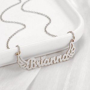 K&ouml;nnte beinhalten: Eine silberne Kettenkette mit einem wei&szlig;en Metallanh&auml;nger, auf dem "Briana" mit Fl&uuml;geln auf beiden Seiten steht.