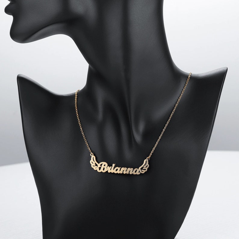 K&ouml;nnte beinhalten: Eine goldene Halskette mit dem Namen "Brianna" in Kursivschrift mit kleinen Fl&uuml;geln auf beiden Seiten des Namens.