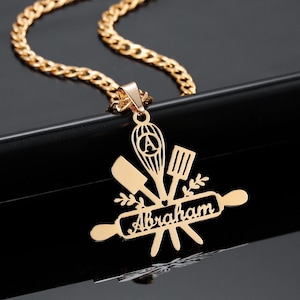 Può includere: Collana d'oro con un ciondolo che presenta un mattarello, una spatola, una frusta e un coltello con il nome "Abraham" sul mattarello.