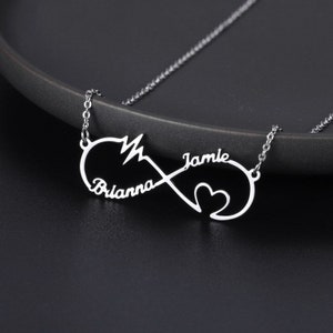 Puede incluir: Un collar plateado con un colgante con forma de símbolo de infinito. El colgante lleva grabados los nombres "Brianna" y "Jamie", junto con un corazón y un símbolo de latido. El collar se muestra sobre un fondo oscuro.