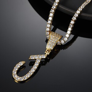 May include: A gold chain necklace with a large, letter J pendant. Both the chain and pendant are encrusted with clear cubic zirconia stones.