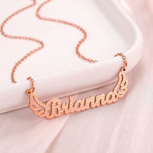 K&ouml;nnte beinhalten: Eine Halskette aus Ros&eacute;gold mit einem Namensschild-Anh&auml;nger, auf dem "Brianna" mit Fl&uuml;geln auf beiden Seiten steht.