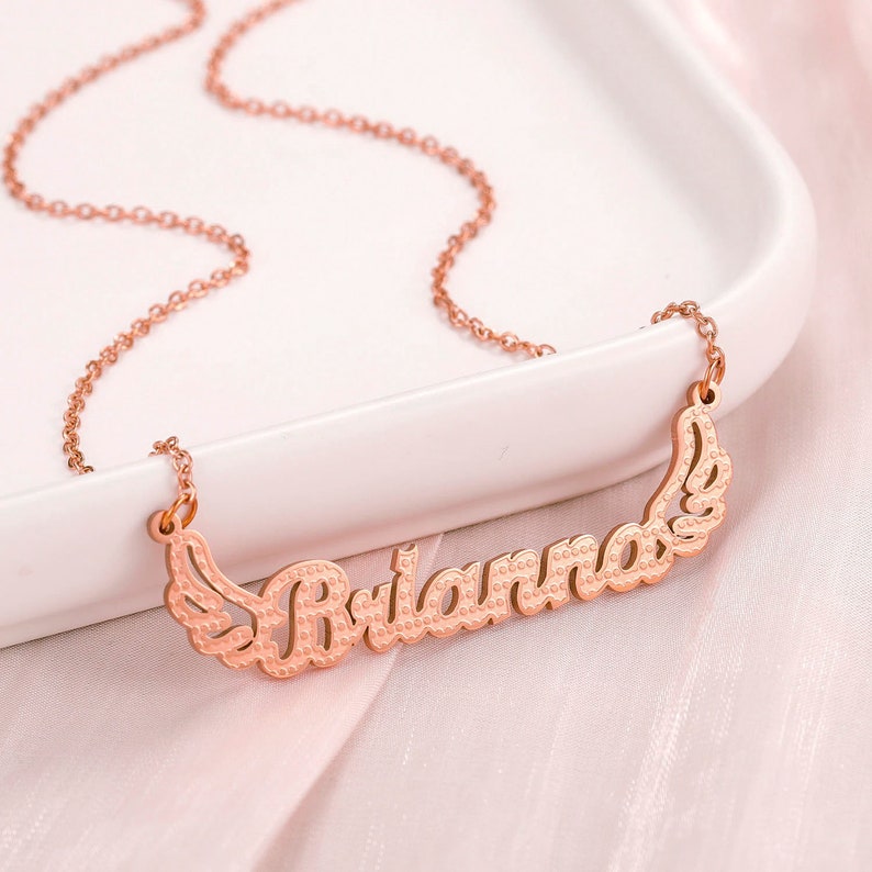 K&ouml;nnte beinhalten: Eine Halskette aus Ros&eacute;gold mit einem Namensschild-Anh&auml;nger, auf dem "Brianna" mit Fl&uuml;geln auf beiden Seiten steht.