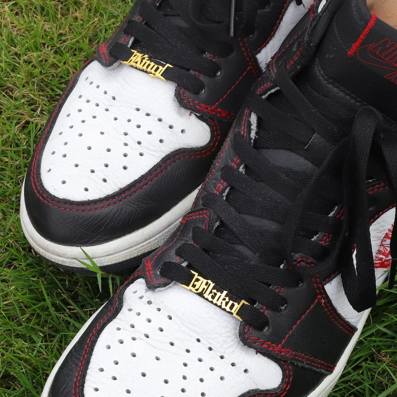 May include: A pair of black and white sneakers with red stitching. The laces are black and have gold charms that say "King" and "Flako".