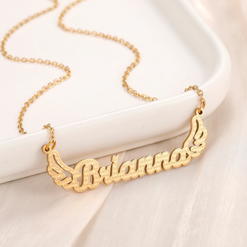 K&ouml;nnte beinhalten: Goldkette mit einem Namensschild-Anh&auml;nger, auf dem "Brianna" mit Fl&uuml;geln auf beiden Seiten steht.
