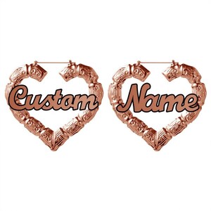May include: A pair of rose gold heart-shaped hoop earrings with the words "Custom" and "Name" written in black cursive lettering.