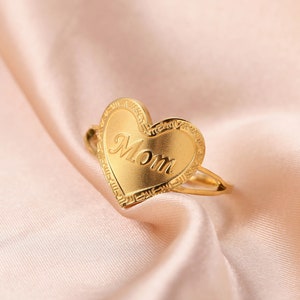 May include: A gold-colored ring with a heart-shaped face. The word "Mom" is inscribed in cursive on the heart. The ring has a decorative border and is set against a soft, pink background.