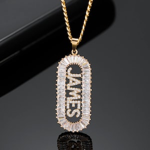 Può includere: Una collana con catena dorata con un ciondolo che presenta il nome "JAMES" scritto in pietre di zircone cubico trasparenti. Il ciondolo è circondato da un bordo di pietre di zircone cubico più piccole.