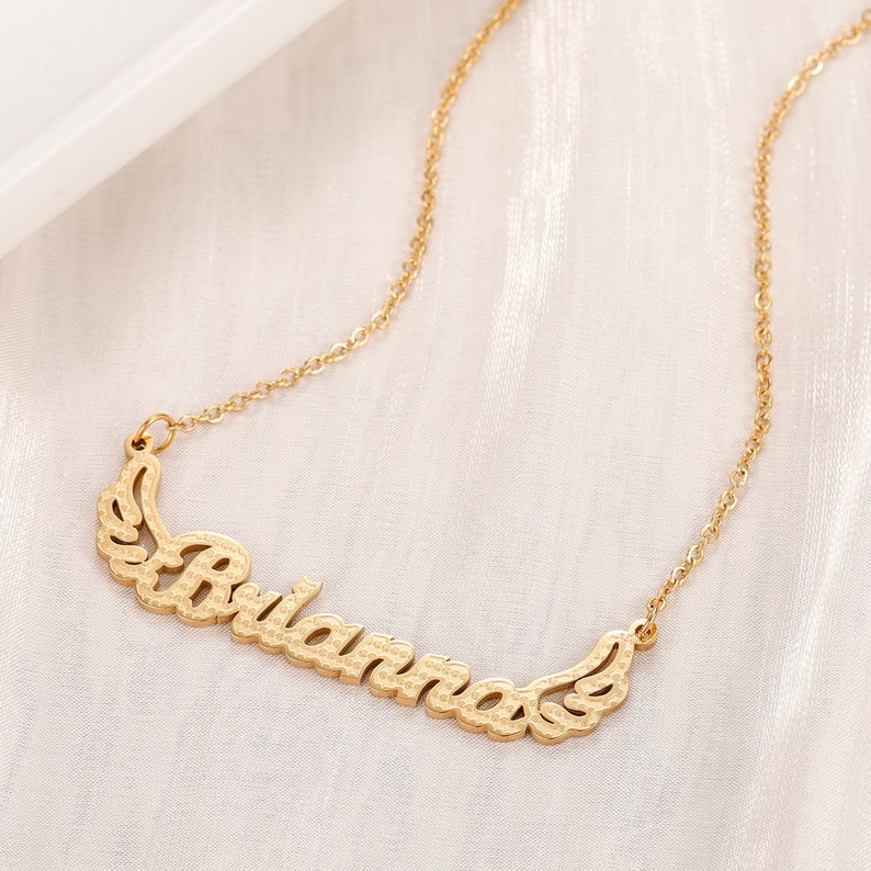 K&ouml;nnte beinhalten: Goldkette mit einer zarten Kette und einem Anh&auml;nger mit dem Namen "Briana" mit Fl&uuml;geln auf beiden Seiten.