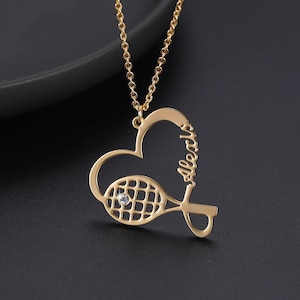 Puede incluir: Collar de raqueta de tenis de oro con un colgante en forma de corazón con la palabra "always" inscrita. La raqueta tiene un pequeño cristal transparente en el centro.