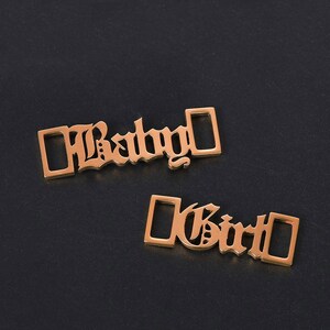 May include: Two gold-colored metal charms with the words "Baby" and "Girl" in a gothic font.