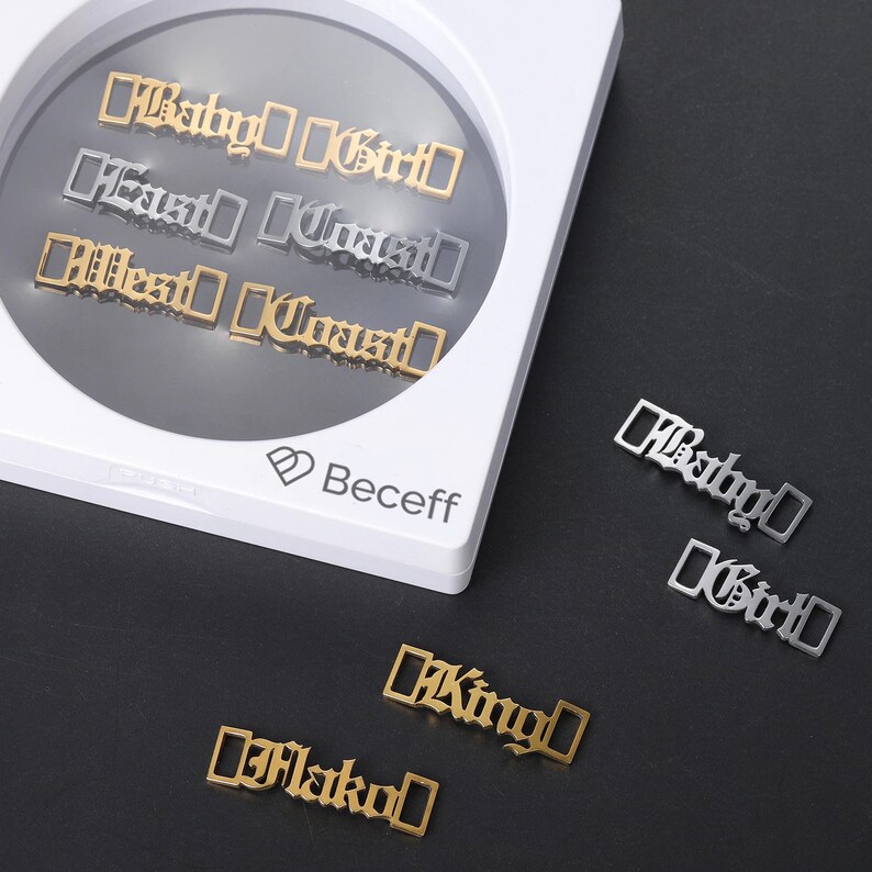May include: A set of gold and silver metal charms with different words, including "Baby Girl", "East Coast", "West Coast", "King", and "Flako". The charms are displayed in a white box with the word "Beceff" printed on it.