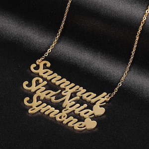 Pode incluir: Colar de ouro com quatro nomes, "Samyra", "Shea", "Nyia" e "Symone", escritos em letra cursiva com um pequeno pingente de coração entre cada nome.