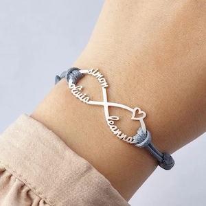 Puede incluir: Una pulsera de plata de infinito con los nombres "Simon", "Olivia" y "Leanna" inscritos en ella. La pulsera está hecha de un cordón gris y tiene un pequeño dije en forma de corazón.
