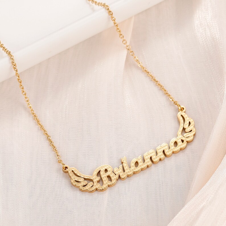 K&ouml;nnte beinhalten: Goldkette mit dem Namen "Brianna" in Schreibschrift mit Fl&uuml;geln auf beiden Seiten.