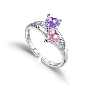 May include: Silver ring with two heart-shaped gemstones, one purple and one pink. The ring has the names "Emma" and "Liana" engraved on it.