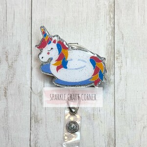 Pode incluir: Um unicórnio branco com uma crina e cauda arco-íris está sentado em um flutuador inflável azul e branco. O unicórnio está usando um porta-crachá de glitter prateado com uma alça de plástico transparente e um botão de glitter prateado.