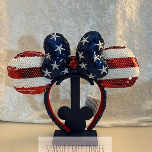 Può includere: Una fascia patriottica con orecchie e fiocco con paillettes. Le orecchie sono a righe rosse e bianche, mentre il fiocco è blu navy con stelle bianche. La fascia è rossa e blu navy, e il testo "SPARKLE CRAFT CORNER" è visibile.