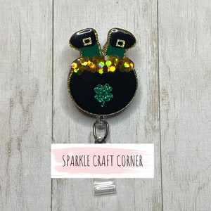 May include: A black pot of gold with green shamrocks and gold glitter, with two green leprechaun legs sticking out of the top. The pot is attached to a clear plastic badge holder with the text "SPARKLE CRAFT CORNER" on a white background.
