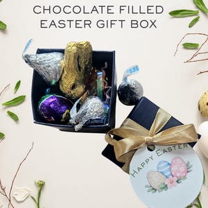 May include: An Easter gift box filled with chocolate candies, including foil-wrapped bunnies and eggs. The box is black with a gold ribbon and a tag that says "Happy Easter" with decorated eggs. The background includes eggs and greenery.