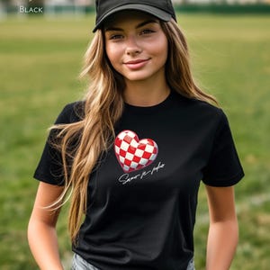 May include: A black t-shirt featuring a red and white chequered heart with the text "Samo je jedno" in white. The model is wearing a black baseball cap and blue jeans.