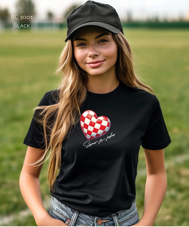 May include: A black t-shirt featuring a red and white chequered heart with the text "Samo je jedno" in white. The model is wearing a black baseball cap and blue jeans.