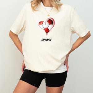 Op de afbeelding: Een wit T-shirt met een rood-witte hartvormige voetbalafbeelding en het woord "KROATI&Euml;" eronder gedrukt. Het model draagt zwarte fietsbroeken.