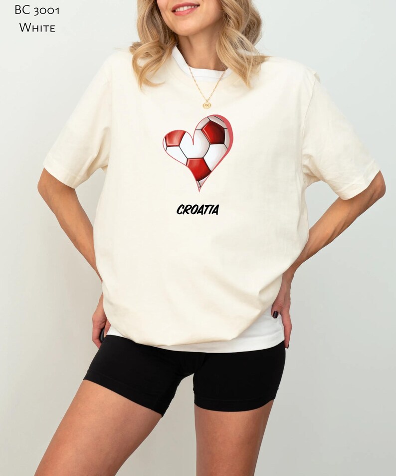 Op de afbeelding: Een wit T-shirt met een rood-witte hartvormige voetbalafbeelding en het woord "KROATI&Euml;" eronder gedrukt. Het model draagt zwarte fietsbroeken.